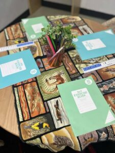 Round table with animal-patterned cloth, four turquoise exercise books, rulers, and a jar of colored pencils. A small plant adds a vibrant touch. Cozy and studious atmosphere.