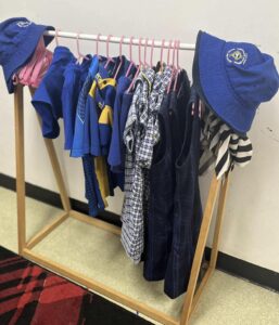 Clothing rack with children's blue and plaid primary school uniforms, hats hanging on wooden frame. Pink hangers add contrast. Organized and tidy display.