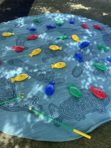 A round mat with fish outlines on the ground displays colorful plastic fish in red, yellow, green, and blue. A toy fishing rod lies nearby, suggesting a playful scene under dappled sunlight.