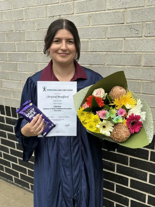 Tillys Gillieston Heights Graduate in a navy gown holds a diploma, a bouquet of colorful flowers, and a chocolate bar, smiling against a brick wall. Celebration and achievement.