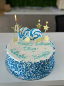 A round birthday cake with blue and white frosting, decorated with two swirl lollipops and candles shaped like stars and a number 4. The blue text on top of the cake reads "Happy Birthday Tillys Gillieston Heights"