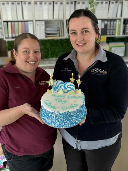 Tillys Gillieston Heights Two Tillys staff members are smiling while holding a cake with blue and white frosting, topped with candles shaped like stars and numbers. Office shelves with binders are in the background.