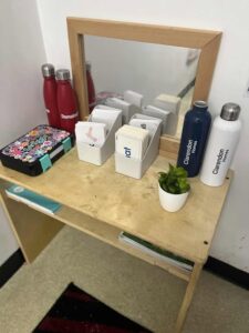A small wooden shelf holds colorful water bottles, floral tin, card holders, and a small potted plant, with a mirror reflecting the items. Cozy and organized.