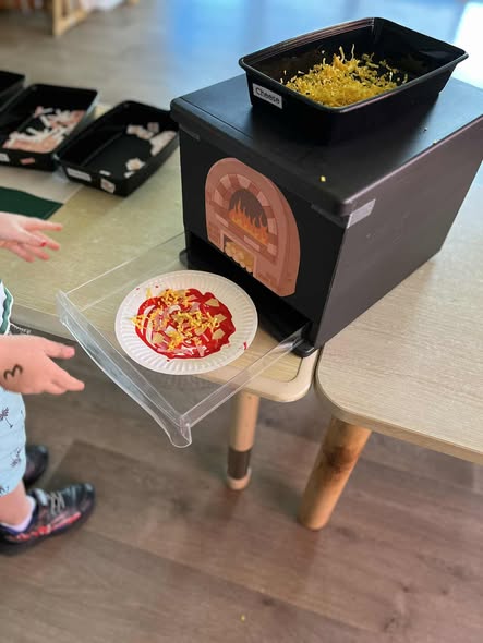 Tillys Rutherford A child, partially visible, interacts with a play pizza station. A toy oven with a pizza paper plate on a tray displays toy cheese and toppings. Playful atmosphere.