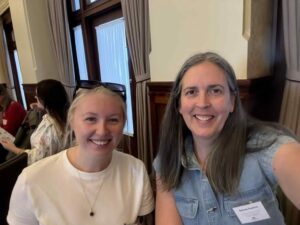 Two women smile at the camera in a room with wooden panels and curtains. One wears a white shirt, the other a denim top with a name tag. The mood is friendly and warm.