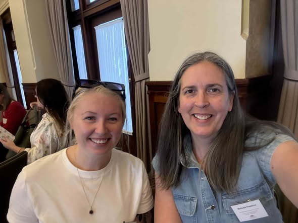 Tillys Rutherford Two women smile at the camera in a room with wooden panels and curtains. One wears a white shirt, the other a denim top with a name tag. The mood is friendly and warm.