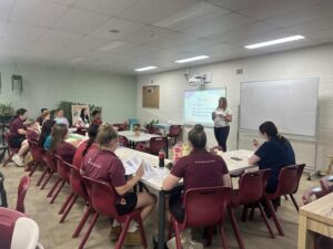 A teacher presents to a classroom of adult students seated at tables with papers and beverages. A slideshow is projected on the wall. The atmosphere is focused.