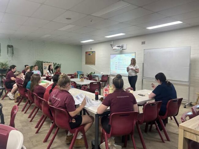 Tillys Waratah A teacher presents to a classroom of adult students seated at tables with papers and beverages. A slideshow is projected on the wall. The atmosphere is focused.