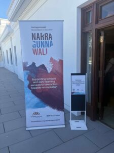 Banner with "Narragunnawali: Reconciliation in Education" and colorful abstract design, stands beside a sign near an open doorway on a sunny day.