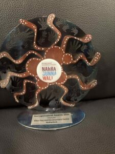 Award plaque with intricate Indigenous design featuring handprints and swirling patterns. Central text: "NAKRA GUNNA WAL'." Tone: celebratory and cultural.