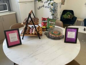 A table with assorted snacks including a tiered stand of mini muffins, bags of Doritos, and a round cookie cake. Two framed notes flank the display. Bright and inviting setting.
