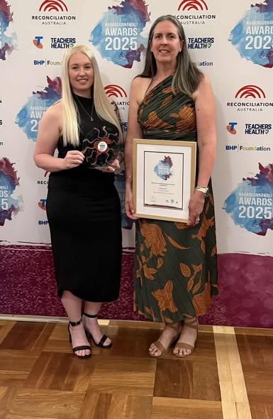 Tillys Rutherford Two women stand smiling at an awards ceremony. One holds a trophy, the other a framed certificate. The backdrop features "Reconciliation Awards 2025" logos.