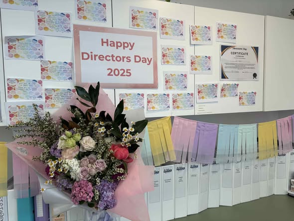 Tillys Cooranbong A wall displays colorful cards and a "Happy Directors Day 2025" sign. Below, a pastel bouquet complements a garland of pastel fringe. Bright, celebratory atmosphere.