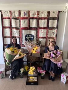 Tillys Waratah 2 Two women sit on wicker chairs holding flower bouquets, with gifts around them. A quilt with "In this together" hangs behind, creating a warm, supportive atmosphere.
