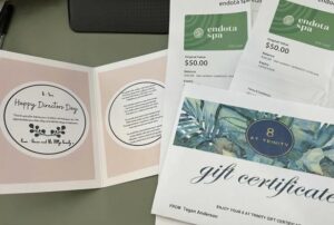 Gift certificates and a card are arranged on a table. The card reads "Happy Directors Day" with a pink floral design. The certificates include Endota Spa and a gift for "8 At Trinity." The mood is celebratory and appreciative.