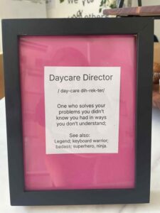 Framed sign with a pink background reading: "Daycare Director: One who solves problems you didn’t know you had." Humorous tone, praising directors.