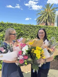 Tillys Waratah 2 Two women smiling outdoors, each holding colorful flower bouquets with pink, yellow, and white blooms. A lush green hedge and palm tree are behind them.