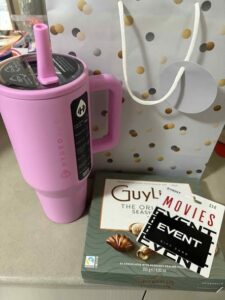 A pink Hydro Jug with a straw, a gift bag with polka dots, Guylian chocolates, and Event Movies gift cards arranged on a countertop.