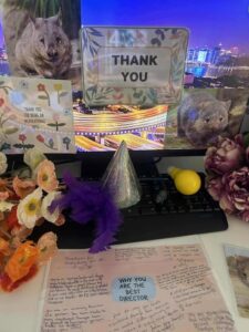 A decorated desk with thank you notes, a "THANK YOU" sign, and a "WHY YOU ARE THE BEST DIRECTOR" poster. Flowers, party hats, and wombat images enhance the celebration.