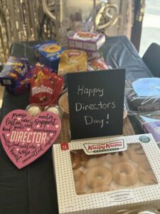 A festive table with snacks, doughnuts, and a heart plaque with words like "praise" and "support." A sign reads "Happy Directors Day!" in a cheerful setting.