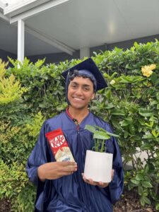 Tillys Waratah 2 A graduate in a navy cap and gown smiles, holding a KitKat bar and a potted plant. They stand in front of lush green bushes, conveying joy and achievement.