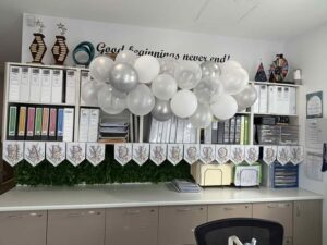 Tillys Gillieston Heights Office decor with white and silver balloons and a banner reading "HAPPY BIRTHDAY." Files and folders line the shelves. The wall says, "Good beginnings never end."