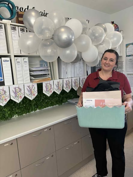 Tillys Gillieston Heights A woman in a red shirt holds a light blue gift basket with balloons floating above. She stands beside a festive banner and a leafy countertop, smiling warmly.