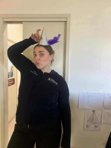 A person wearing a playful expression sports a cone-shaped party hat with purple feathers. They stand in a doorway, with children’s drawings on the wall nearby.