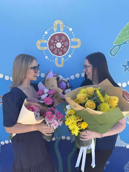 Tillys Waratah 2 Two women smiling at each other hold vibrant flower bouquets against a blue mural backdrop, conveying a joyful and celebratory mood.