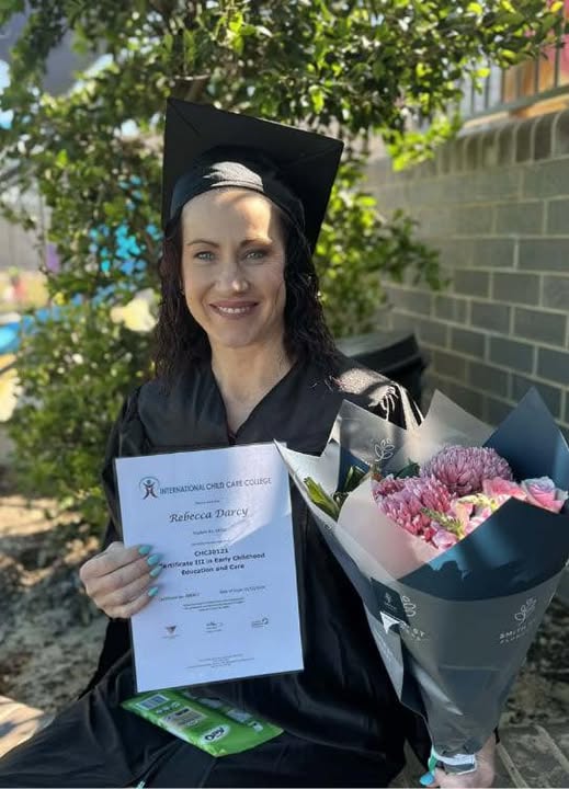 Tillys Chisholm Graduate in cap and gown smiles, holding a certificate and a bouquet of flowers, seated outdoors against a backdrop of greenery and a brick wall.