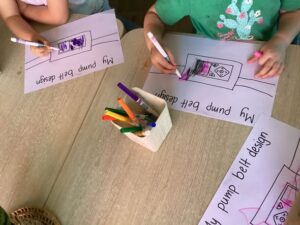 Children are sitting around a table, coloring on papers titled "My pump belt design." A cup with colorful markers is in the center, creating a playful and creative mood.