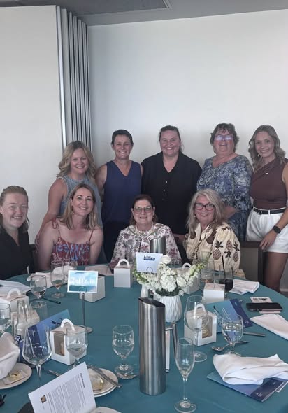 A group of nine people at a banquet table, smiling for a photo. The table has teal cloth, white flowers, and place settings, creating a joyful atmosphere.