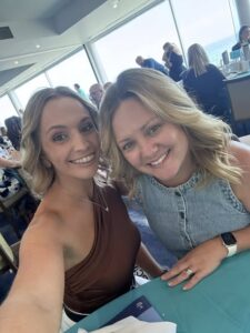 Two women smiling for a selfie in a bright, busy restaurant with ocean views. They appear relaxed and happy, wearing casual summer attire.