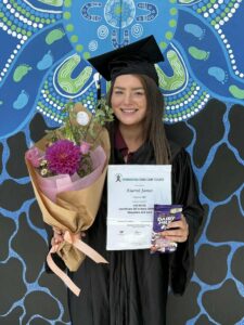 Graduating student in a cap and gown holds a certificate, a bouquet with pink flowers, and a chocolate bar. Background features a colorful abstract mural.