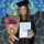 Graduating student in a cap and gown holds a certificate, a bouquet with pink flowers, and a chocolate bar. Background features a colorful abstract mural.