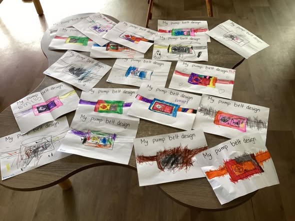 Several colorful children's drawings of "My pump belt design" are spread on a wooden table. The mood is creative and playful.