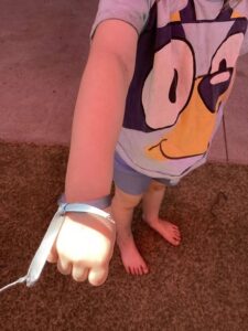A child stands on grass, extending an arm with a bracelet tied around the wrist. Their shirt features a cartoon face with a playful expression.
