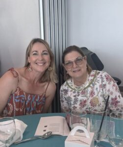 Two smiling women sit at a teal-colored table. One wears a patterned sundress, and the other a floral top with glasses. The atmosphere is cheerful and relaxed.