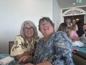 Two women are seated at a table, smiling warmly. They are surrounded by others in a lively indoor setting with soft lighting, creating a happy and relaxed atmosphere.