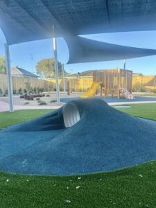 Playground with a grassy tunnel slide, shaded by a large canopy. In the background, a wooden climbing structure with a yellow slide. Peaceful atmosphere.