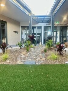 Outdoor courtyard with a small rock-lined dry creek bed, surrounded by green plants and mulch. Modern building entrance in the background under a glass roof, creates a welcoming and serene atmosphere.