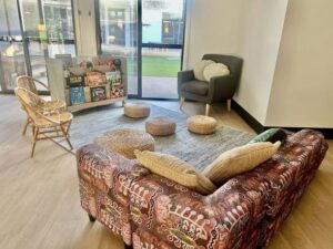 Cozy reading nook with a colorful patterned couch, wicker chairs, and poufs on a rug. Books are displayed near a window with a garden view.