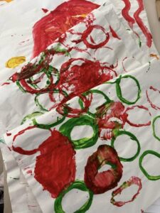 Tillys Rutherford A wicker basket holds Christmas wrapping paper with red and green circle patterns. Beside it, a small seasonal floral arrangement and a sign inviting people to take one. The background features patterned wall tiles.