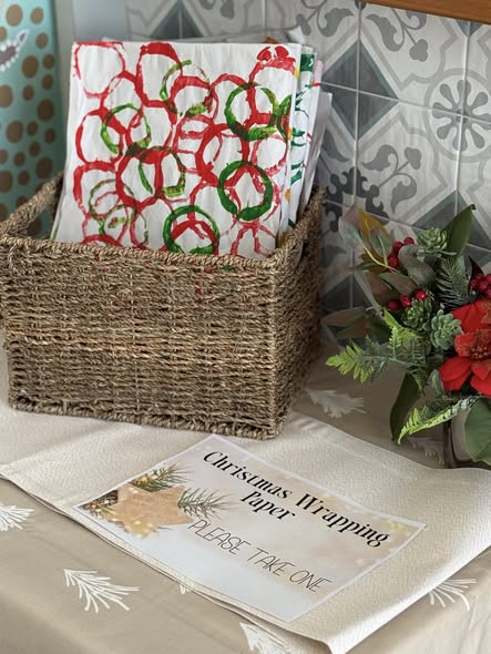 A wicker basket holds Christmas wrapping paper with red and green circle patterns. Beside it, a small seasonal floral arrangement and a sign inviting people to take one. The background features patterned wall tiles.