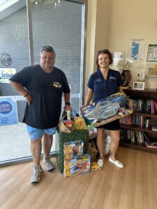A man and woman smile while holding toys and a gift bag in a room with bookshelves. The scene conveys a cheerful and charitable atmosphere.