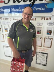 Man smiling, wearing a dark shirt with neon accents, holding a festive gift bag in front of a wall with educational posters and documents.