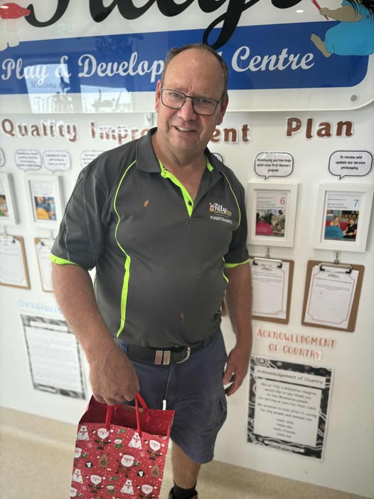 Man smiling, wearing a dark shirt with neon accents, holding a festive gift bag in front of a wall with educational posters and documents.