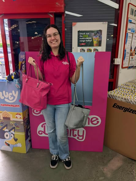 A smiling person in a pink shirt holds a pink and a gray handbag in front of a pink display. Colorful boxes and posters are in the background.