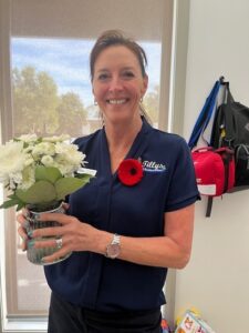 A smiling woman in a navy shirt holds a vase of white flowers. She wears a red poppy pin. Behind, bags hang on a white wall. Bright and cheerful atmosphere.