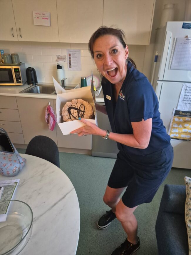 Tillys Fern Bay A joyful woman in navy attire holds a cake box in a kitchen, smiling widely. The kitchen has a fridge, table, and assorted kitchen items.